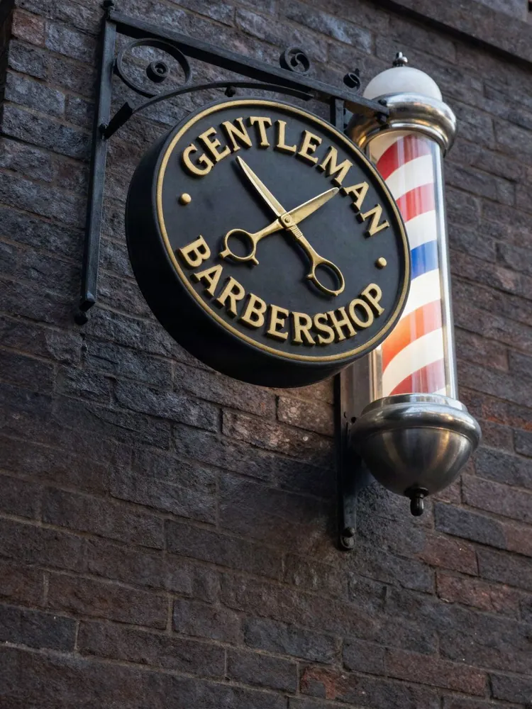 벽돌 벽에 달린 둥근 검은 돌출 간판 시안. 금색으로 GENTLEMAN BARBERSHOP 글자와 가위가 새겨져 있고 옆에 이발소 기둥이 있습니다